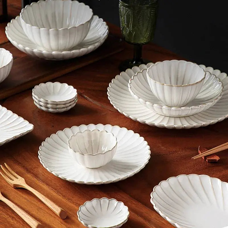 Full ceramic tableware set with scalloped white bowls and plates arranged on a wooden table. Durable and stylish dining essential collection.