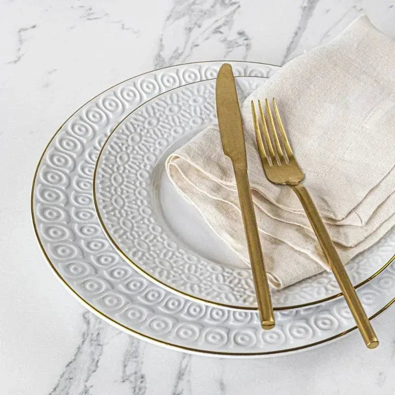 Close-up of Sofia Vergara's White Stoneware Set plate with gold trim, paired with gold flatware and a linen napkin for a refined table setting.