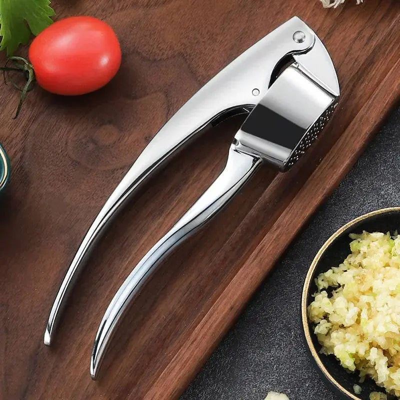 Shiny Stainless Steel Garlic Masher - Manual Garlic Crusher on a wooden surface with minced garlic, tomato, and herbs around.