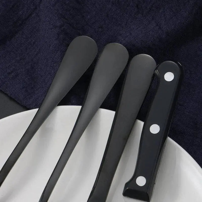 24 Pieces Stainless Steel Cutlery Set | Durable Dining Elegance displaying the sleek black handles of spoons and a steak knife on a white plate.