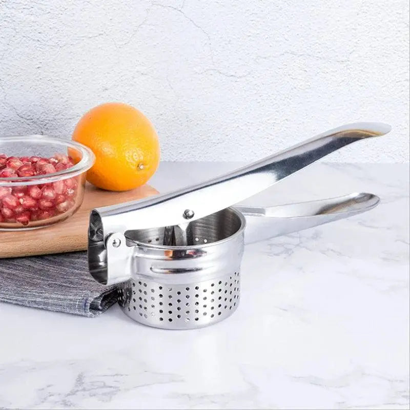 Stainless Steel Manual Juicer - Multifunctional | Compact Design placed on a kitchen counter next to an orange and pomegranate seeds.
