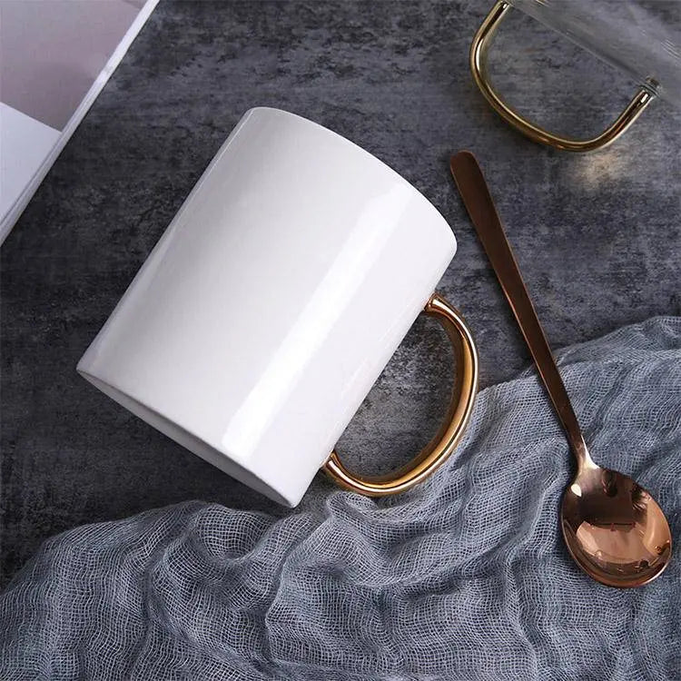 Elegant Coffee Cup with Gold Handle lying on a gray cloth with a matching gold spoon, emphasizing its chic and modern appeal.