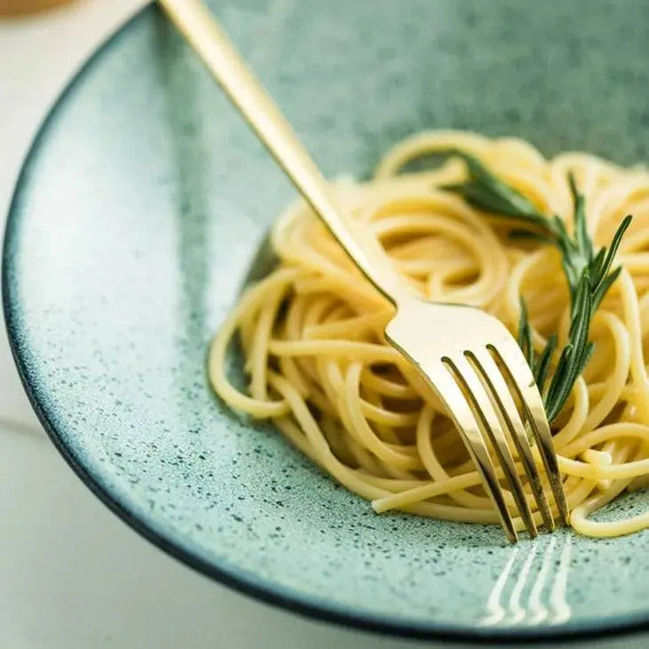 Golden fork from the 24-piece cutlery set twirling spaghetti in a green ceramic bowl, perfect for festive meals.